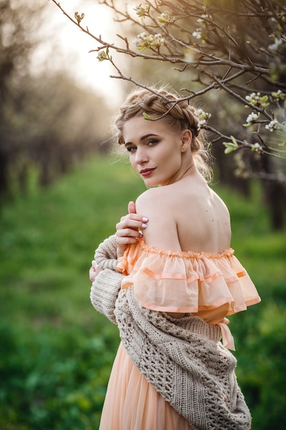 Menina com cabelo loiro em um vestido leve no jardim florido. conceito de moda feminina primavera. garota em um lindo vestido e suéter de malha aprecia o pôr do sol em um jardim florido de pera