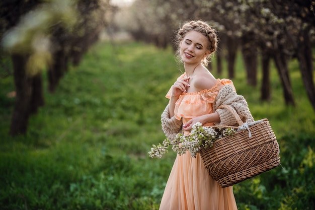 Menina com cabelo loiro em um vestido leve no jardim florido. garota em um lindo vestido e suéter de tricô aprecia o pôr do sol em um jardim florido de pera, com uma cesta de flores