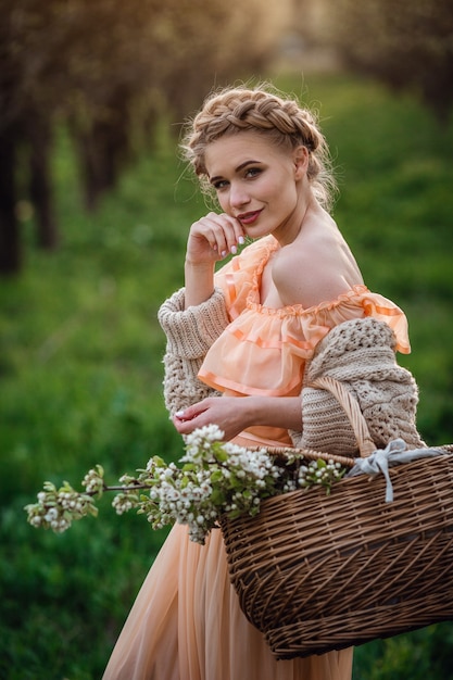 Menina com cabelo loiro em um vestido leve no jardim florido. garota em um lindo vestido e suéter de tricô aprecia o pôr do sol em um jardim florido de pera, com uma cesta de flores