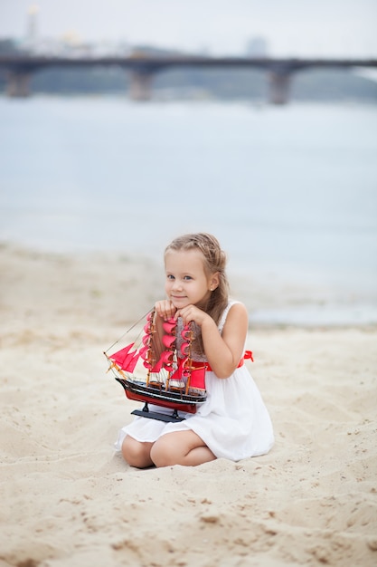 Menina com cabelo loiro no vestido branco, segurando o navio com velas escarlate. Criança sente-se à beira-mar, tem um veleiro de brinquedo nas mãos.