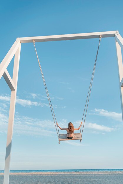 Menina com cabelos longos em corda balança contra o mar e fundo de céu azul Conceito de liberdade e descuido infantil Quadro vertical