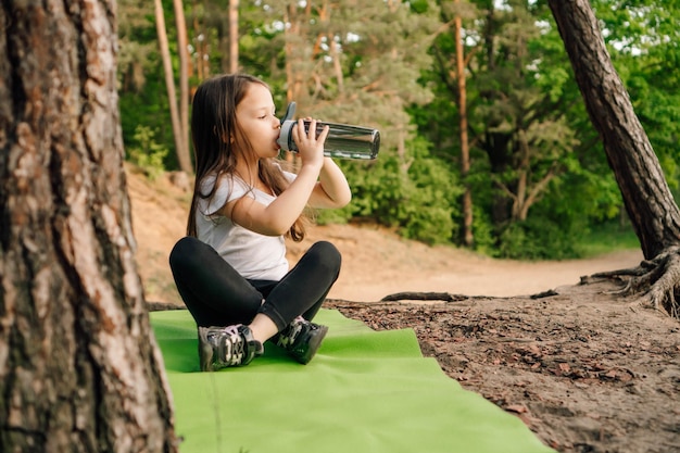 Foto menina com cabelos longos escuros sentada no tapete verde na natureza e bebendo água da garrafa descansando depois do treino