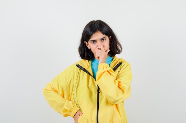 Menina com camisa, jaqueta, segurando a mão na boca enquanto mantém a mão no quadril e olhando pensativa, vista frontal.