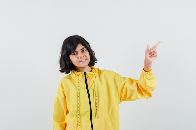 Menina com capuz amarelo apontando para o canto superior direito e parecendo confiante, vista frontal.