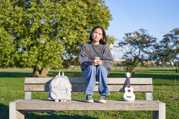 Menina com cara triste sentada no banco com smartphone e ukulele parecendo chateada e decepcionada por estar