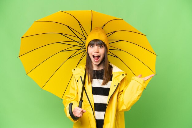Menina com casaco à prova de chuva e guarda-chuva sobre fundo croma chave isolado com expressão facial chocada