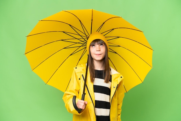 Menina com casaco à prova de chuva e guarda-chuva sobre fundo croma chave isolado e olhando para cima