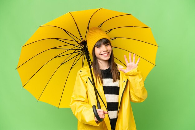 Menina com casaco à prova de chuva e guarda-chuva sobre fundo croma isolado contando cinco com os dedos