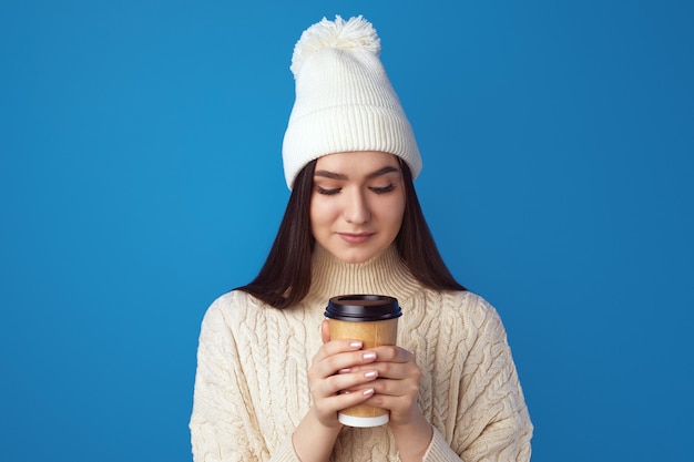 Menina com chapéu branco e suéter gosta de beber café em uma xícara de comida para viagem