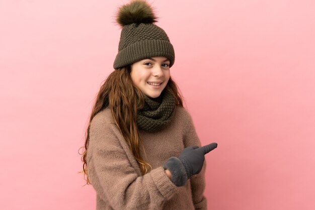 Menina com chapéu de inverno isolada em fundo rosa apontando para trás