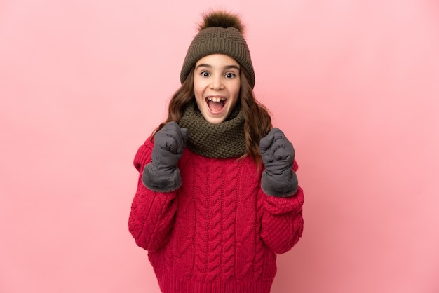 Menina com chapéu de inverno isolada em fundo rosa comemorando vitória na posição de vencedora