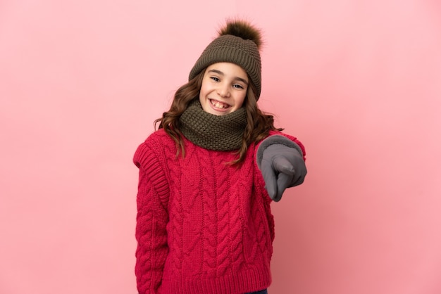 Menina com chapéu de inverno isolada em um fundo rosa apontando para a frente com uma expressão feliz