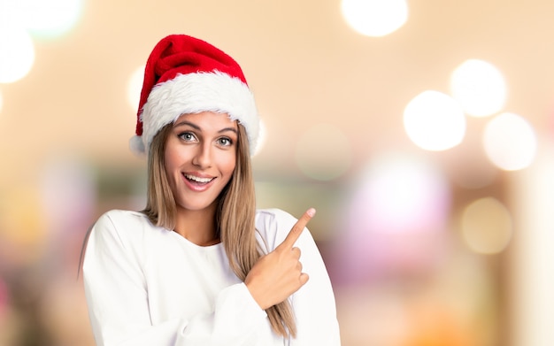 Menina com chapéu de Natal, apontando o dedo para o lado sobre parede fora de foco