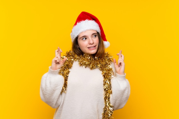 Menina com chapéu de Natal isolado muro amarelo com dedos cruzando