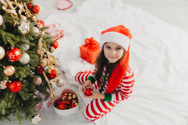 Menina com chapéu de Papai Noel decora a árvore com brinquedos. Um lindo bebê está se preparando para celebrar o Natal.