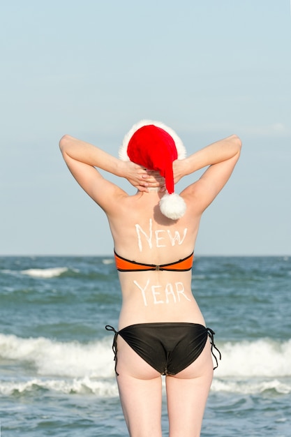 Menina com chapéu de Papai Noel e maiô fica na praia. Mãos atrás da cabeça. Inscrição de ano novo na parte de trás