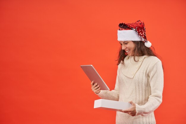 Menina com chapéu de Papai Noel feliz com seu novo presente para tablet em fundo laranja