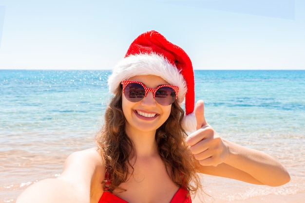 Menina com chapéu de Papai Noel mostrando o polegar para cima e se divertindo tirando fotos de selfie no smartphone