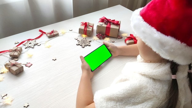 Menina com chapéu de Papai Noel segurando o smartphone com tela verde sobre fundo branco de mesa de madeira com presentes de Natal.