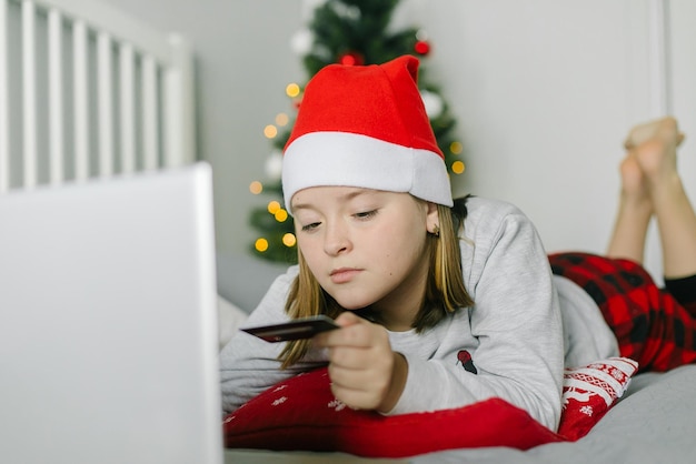 Foto menina com chapéu de papai noel vermelho segurando o carrinho de crédito, usando o laptop do dispositivo. conceito de compras online
