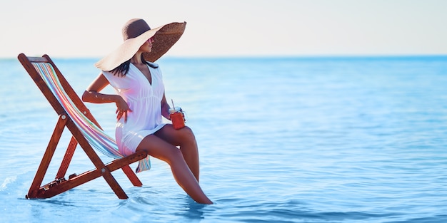 Menina com chapéu de sol e túnica sentada na cadeira de praia à beira-mar enquanto dá coquetéis de férias de verão