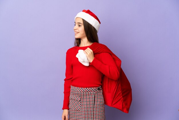 Menina com chapéu e saco de Natal isolada na parede roxa olhando para o lado e sorrindo