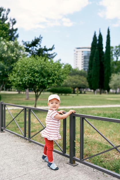 Menina com chapéu-panamá parada perto de uma cerca de metal em um parque verde