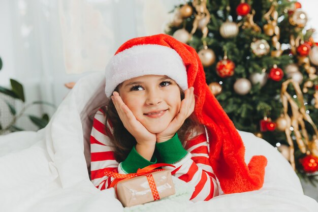 Menina com chapéu vermelho de ano novo com um presente de Natal. Ela está deitada na cama, abraçando uma caixa contra o fundo de uma árvore de Natal.