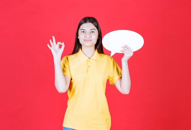 Menina com dresscode amarelo segurando um quadro oval de informações e mostrando um sinal positivo com a mão