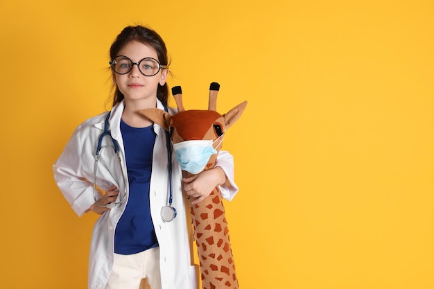 Menina com estetoscópio de óculos vestida de médico abraçando girafa de brinquedo no espaço de fundo amarelo para prática de pediatra de texto