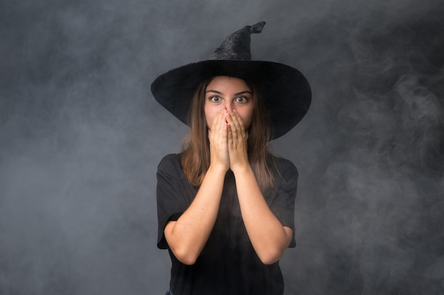 Foto menina com fantasia de bruxa para festas de halloween ao longo da parede escura isolada com expressão facial de surpresa