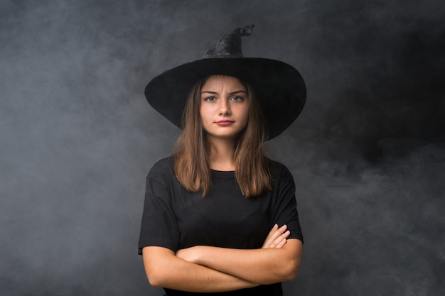 Menina com fantasia de bruxa para festas de halloween ao longo da parede escura isolada, mantendo os braços cruzados