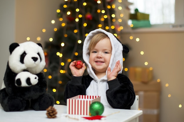 Menina com fantasia de panda e o brinquedo de panda abrindo um presente de Natal embaixo da árvore de Natal