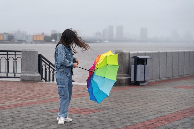 Menina com guarda-chuva de arco-íris na margem em um dia chuvoso e ventoso