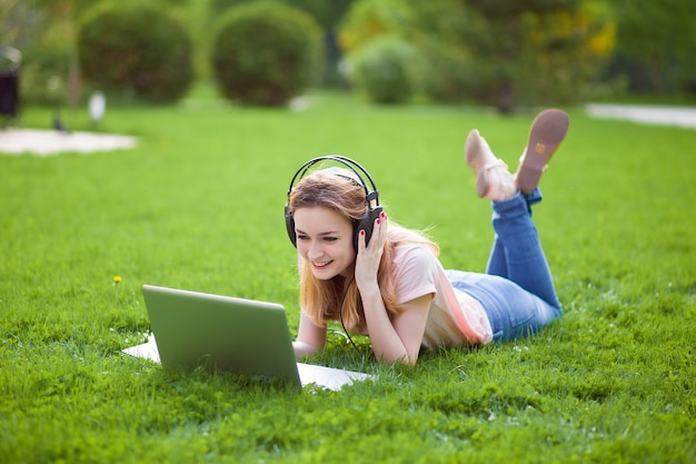 Menina com laptop em fones de ouvido deitada na grama