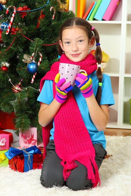 Menina com lenço rosa e um copo de bebida quente sentada perto da árvore de Natal