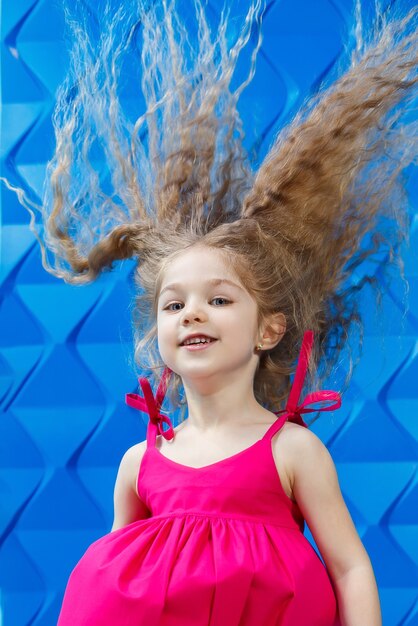 Menina com longos cabelos cacheados em um vestido rosa sobre um fundo azul dança e ri, as emoções das crianças brilhantes de alegria, infância feliz