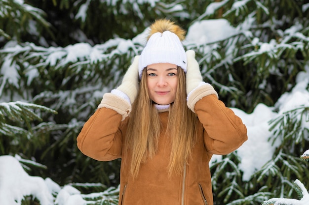 Menina com luvas de chapéu de malha branca em dia de inverno nevado, perto de árvore de natal. jovem sorrindo, brincando com a neve. conceito de inverno