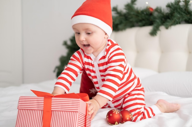 Menina com macacão listrado e chapéu de Papai Noel brincando com uma caixa de presente no quarto decorado de natal