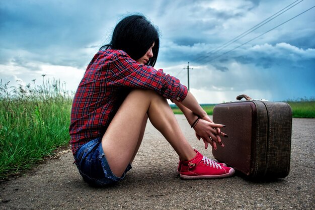 Menina com mala vintage senta-se no carro esperando asfalto. garota viajando pedindo carona.