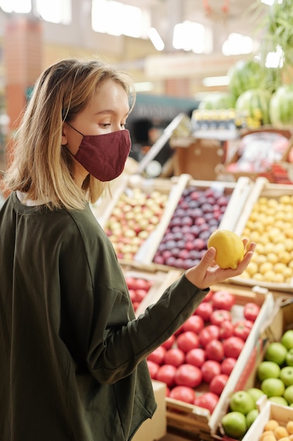 Menina com máscara facial em pé no balcão de frutas e olhando para o limão ao escolher no mercado