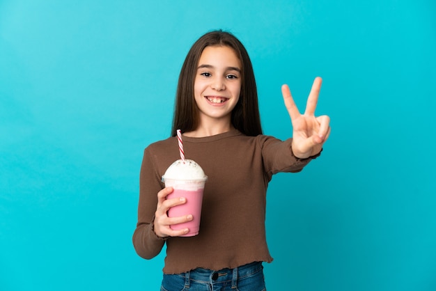 Menina com milk-shake de morango isolada na parede azul sorrindo e mostrando sinal de vitória