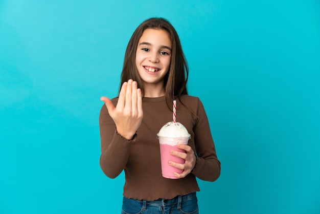 Menina com milk-shake de morango isolado sobre fundo azul, convidando para vir com a mão. Feliz que você veio