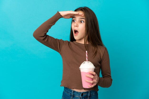 Menina com milkshake de morango isolada em um fundo azul fazendo gesto surpresa enquanto olha para o lado