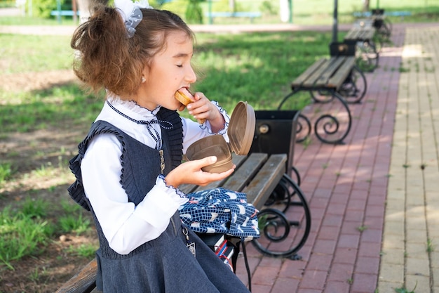 Menina com mochila comendo sanduíche embalado em uma caixa de sanduíche perto da escola Um lanche rápido com um pão comida não saudável almoço da escola Volta às aulas Educação aulas do ensino fundamental 1º de setembro