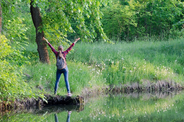 Menina com mochila fica de costas com as mãos contra a floresta e o lago