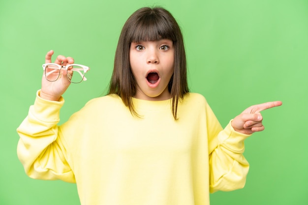 Menina com óculos sobre fundo croma chave isolado surpreso e apontando o lado