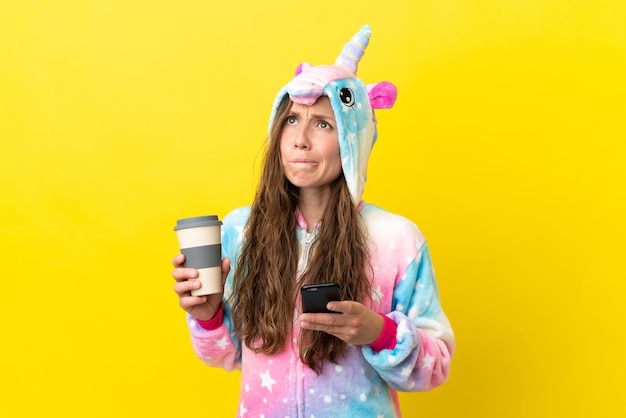 Menina com pijama de unicórnio sobre um fundo isolado segurando um café para levar e um celular enquanto pensa em algo