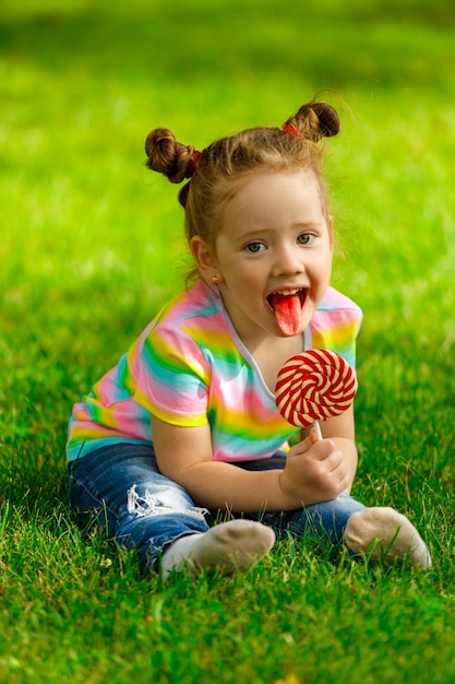 Menina com pirulito vermelho senta-se na grama de verão no parque.