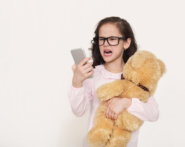 Menina com raiva de óculos com smartphone e ursinho de pelúcia no fundo branco do estúdio, copie o espaço